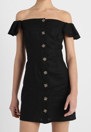 Sort off-shoulder kjole med korte flæseærmer og en række store brune knapper foran, båret af en person, der står.