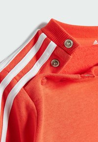 Top sportivo rosso con strisce bianche sulle maniche e bottoni a pressione sul colletto. Tessuto morbido con dettaglio a costine sul collo.