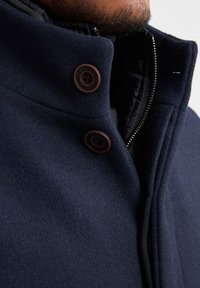 Navyfarbener Wollmischungs-Mantel mit hohem Kragen, ausgestattet mit zwei braunen Knöpfen, einem Reißverschluss und einer glatten Textur.