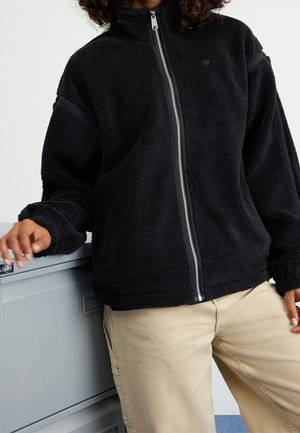 Osoba ubrana w czarną polarową kurtkę na zamek błyskawiczny z beżowymi spodniami, opierająca się o szarą szafkę na akta, z jedną ręką na niej spoczywającą.