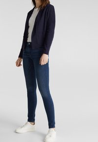 Kvinna klädd i en marinblå kavaj, vit skjorta, mörkblå skinny jeans och vita sneakers, stående mot en enfärgad bakgrund.