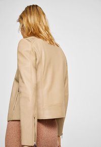 Žena s vlasem po ramena v blond barvě, nosící béžovoukoženou bundu a růžovou manšestrovou sukni, stojící zády k jasnému pozadí.