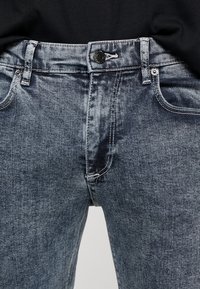 Vaqueros de mezclilla gris oscuro con un diseño clásico de cinco bolsillos, que presentan un cierre de botón y cremallera, y detalles de costura en contraste.