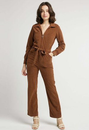 Salopette en velours côtelé brun avec des manches longues, fermeture éclair à l'avant, deux poches poitrine et une ceinture à la taille. Jambes évasées et détail de tissu texturé.