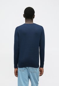 Άνδρας με κοντά σγουρά μαλλιά, που φοράει μπλε μακρυμάνικο πουλόβερ και ανοιχτόχρωμο μπλε τζιν, γυρισμένος μακριά σε άσπρο φόντο.