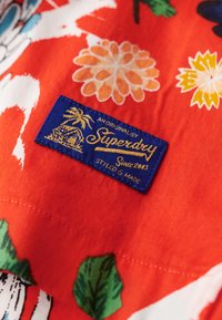 Etiqueta de tela azul que dice "Superdry Since 2003" cosida en una prenda de patrón floral rojo con flores naranjas, amarillas, verdes y blancas.