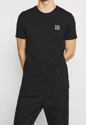Man draagt een eenvoudige zwarte korte mouwen T-shirt met de tekst "MARC O'POLO DENIM" op de linkerborst, gecombineerd met een zwarte broek.