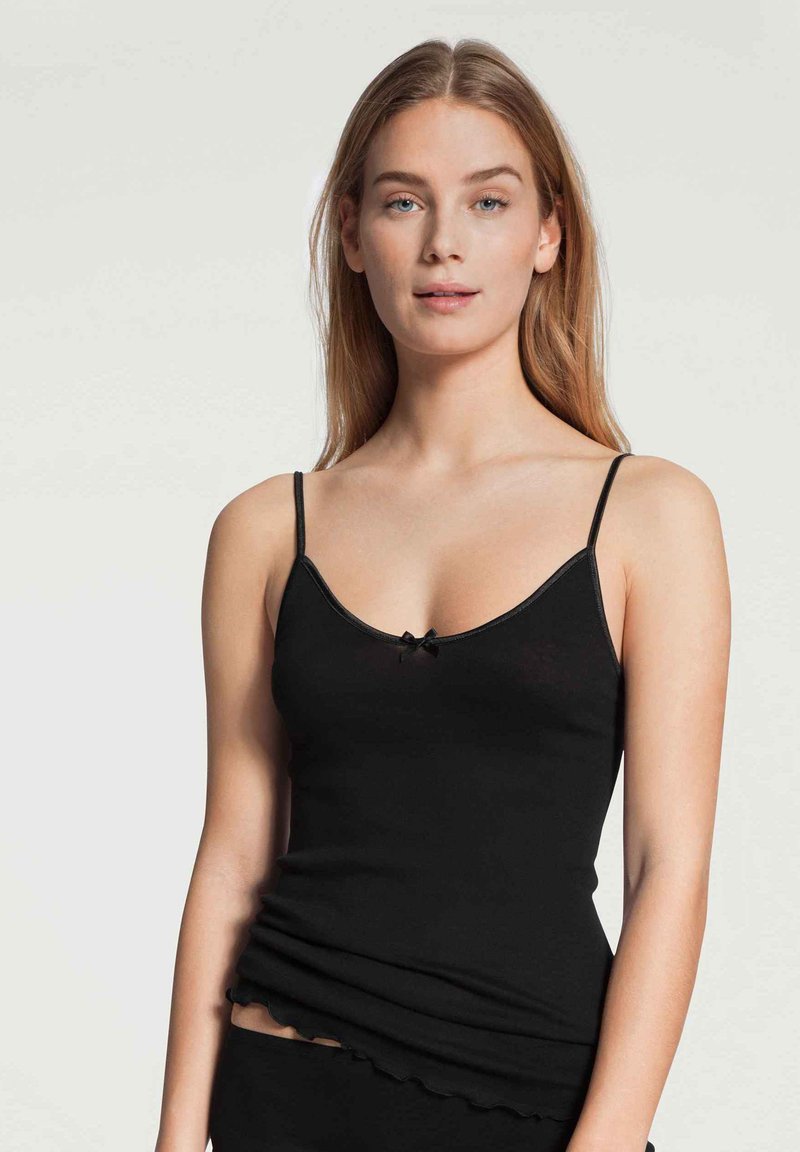 Camisola nera con spalline sottili, scollo a barca e un piccolo dettaglio a fiocco al centro. Presenta un tessuto morbido e testurizzato e un orlo ondulato.