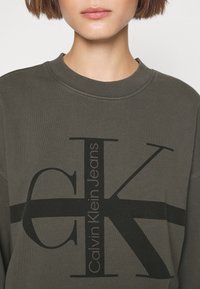 Sudadera verde oliva con cuello redondo que presenta un logo negro de CK y el texto "Calvin Klein Jeans" en la parte frontal. Material de mezcla de algodón.