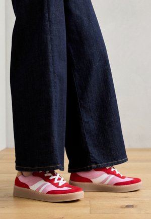 Rode en roze suède sneakers met witte strepen, voorzien van een plat rubberen zool en een vetersluiting. Draagt in combinatie met wijde donkerdenim broeken.