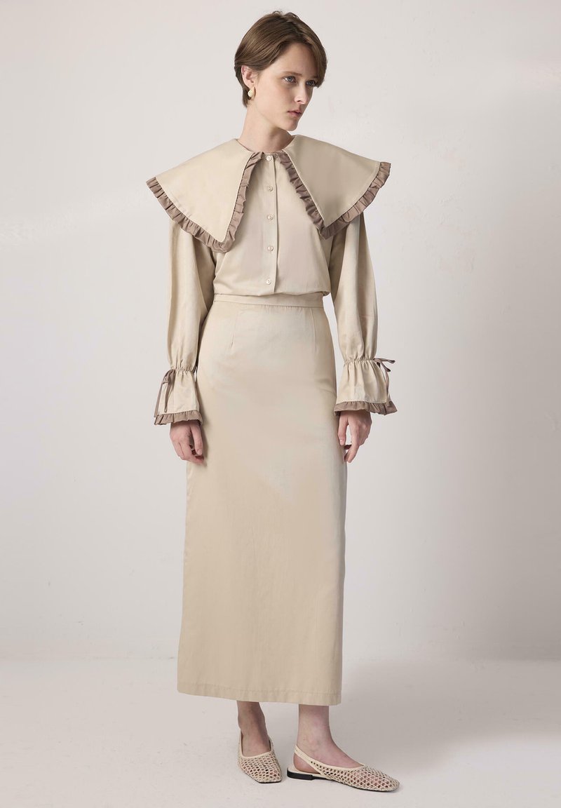 Beige blus med överdimensionerad, volangkrage och knappstängning, uppmatchad med en lång, matchande kjol. Accentdetaljer på ärmarna. Enkel, slät textur.