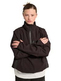 Jeune personne à la peau claire et aux cheveux attachés en arrière, portant une veste noire zippée, bras croisés, faisant face vers l'avant sur un fond blanc.