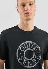 Czarny bawełniany t-shirt z szarą, postarzaną grafiką w kształcie koła z napisem "QUITE SENSE" na środku, okrągły dekolt, krótkie rękawy.