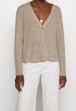 Cardigan en tricot beige avec un col en V, des manches longues et une fermeture à bouton unique, associé à un pantalon large blanc. Le détail du tissu texturé est visible.