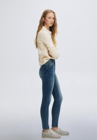 Donna in piedi con le braccia incrociate che indossa un maglione color crema, jeans skinny blu a vita alta e sneakers bianche su uno sfondo chiaro uniforme.