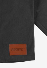 Czarna tkanina o teksturowanej powierzchni z brązową skórzaną metką wytłoczoną napisem "PROSTO" w dolnym lewym rogu.