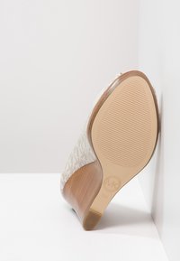 Beige lodička na vysokém podpatku s texturovanou podrážkou opřená o bílou zeď, na podrážce je viditelné logo MK a velikost 5M.