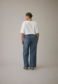White oversized t-shirt with short sleeves, paired with high-waisted, wide-leg blue jeans. Back view shows two rear pockets and a straight silhouette.