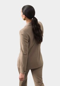 Blazer beige a medida con hombros estructurados, tejido suave y sutiles detalles de costura, llevado sobre unos pantalones a juego.