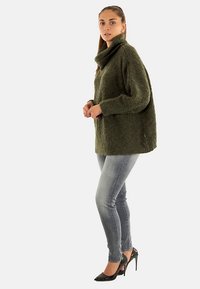 Groen oversized gebreide trui met een hoge col, gestructureerde stof en vallende schouders, gecombineerd met slim-fit blauwe jeans en zwarte hakken.
