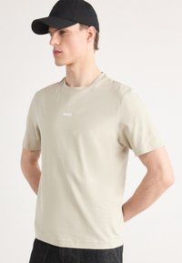 Νεαρός άνδρας με μπεζ μπλουζάκι BOSS και μαύρο καπέλο, στέκεται με τα χέρια πίσω από την πλάτη, κοιτάζοντας αριστερά μπροστά σε απλό λευκό φόντο.