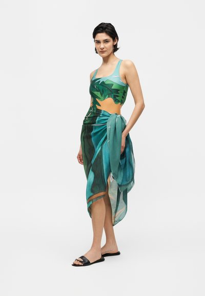 Femme portant une robe bleu-vert à imprimé tropical avec des découpes, nouée à la taille, associée à des sandales noires, debout sur un fond blanc.