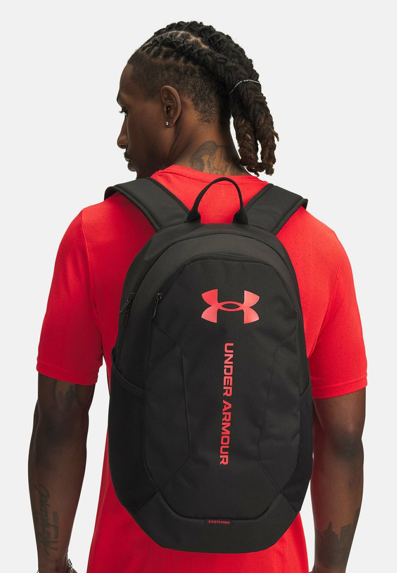 Sac à dos noir avec des accents rouges, présentant le logo Under Armour, un design incurvé et une poche avant. Le matériau semble durable et résistant aux intempéries.