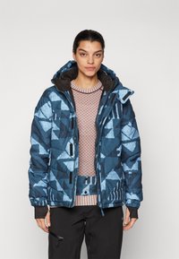Giacca blu trapuntata con motivi geometrici, cappuccio regolabile e dettagli neri. Indossata sopra un maglione rosa chiaro e pantaloni scuri.
