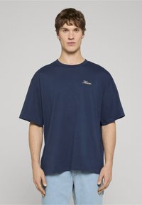 Džinsinė mėlyna medvilninė marškinėliai su laisvu siluetu, trumpomis rankovėmis ir apvalia iškirpte, ant krūtinės yra mažas baltas parašytas logotipas.