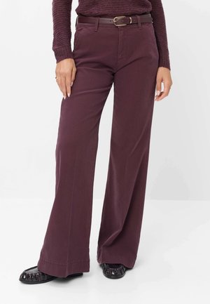 Vrouw draagt bordeaux wijde broek met bijpassende riem, een getextureerde top met lange mouwen en zwarte loafers op een witte achtergrond.