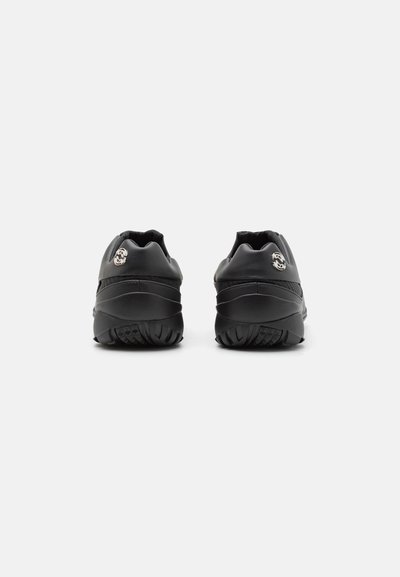 Des chaussures de sport noires en matériau synthétique lisse, avec un talon arrondi, une semelle texturée et un accent métallique sur le côté.