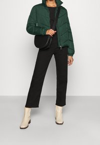 Giacca a vento verde con zip frontale, borsa rotonda a tracolla nera, pantaloni neri a gamba dritta e stivaletti color crema. Tessuto liscio, texture gonfia.