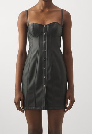 Kobieta ubrana w czarną mini sukienkę z ekologicznej skóry na cienkich ramiączkach, z guziczkami w kształcie serca na przodzie, stojąca na tle jednolitego tła.