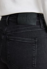 Fekete farmernadrág finom textúrával, látható hátsó zsebbel és Calvin Klein bőrcímkével a deréknál.