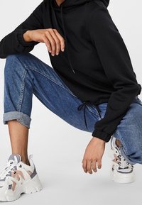 Schwarzer Kapuzenpullover mit elastischen Bündchen, kombiniert mit hochgekrempelten blauen Jeans und klobigen weißen Sneakers mit grauen und rosa Akzenten.