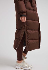 Bruine pufferjas met een lang, gestikt ontwerp, voorzien van zijdrukknoopsluitingen en een contrasterende zwarte binnenvoering. Draag over een paar bruine legging en zilveren sneakers.