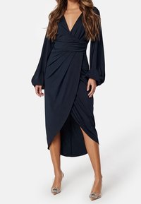 Navy wrap dress with long sleeves, deep V-neck, crossover front, and a side slit. Smooth texture with draping details, paired with embellished heels.
