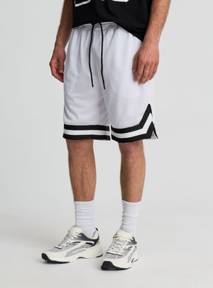 Uomo che indossa pantaloncini da basket in rete bianchi con strisce a chevron nere, calzini bianchi e scarpe sportive bianche e nere, in piedi su pavimento grigio.