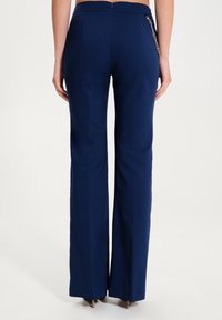 Osoba v námořnicky modrých vysokých širokých kalhotách s zlatým řetízkovým detailem na pravé straně, zobrazená ze zadní strany.