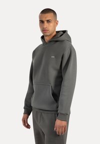 Sudadera gris con un bolsillo frontal, capucha ajustable y un pequeño parche de logo en el pecho. Fabricada con un tejido suave y texturizado.