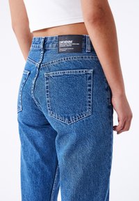 Blue denim jeans with a high waist. Features include two back pockets, contrast stitching, and a branded black label on the waistband.