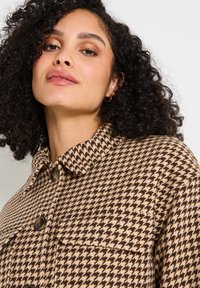 Veste à motif pied-de-poule marron et beige avec un col, fermeture à bouton et poche avant, fabriquée en tissu texturé.