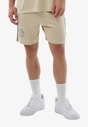 Uomo che indossa pantaloncini beige con toppa "New York", calzini bianchi e sneakers Nike bianche, in piedi su uno sfondo semplice.