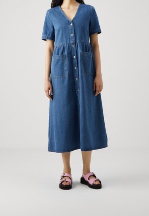 Kvinde iført en blå denim midi-kjole med knapper foran og store lommer, kombineret med lyserøde sandaler med spænder mod en lys baggrund.