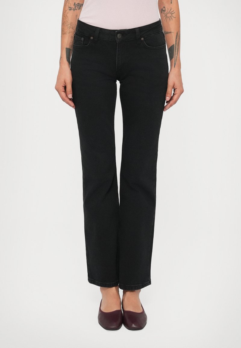 Person iført tætsiddende sorte jeans, lyserød top og mørkerøde flade sko, stående mod en ensfarvet baggrund.