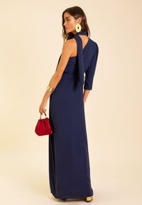Robe longue bleu marine avec des manches trois-quarts et un dos drapé, accompagnée d'un sac à main rouge et de grandes boucles d'oreilles circulaires.