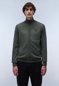 Grön zip-up tröja med hög krage, texturerad övre del och en cirkulär logopatch. Ribbad midja och ärmslut, buren över mörka byxor.