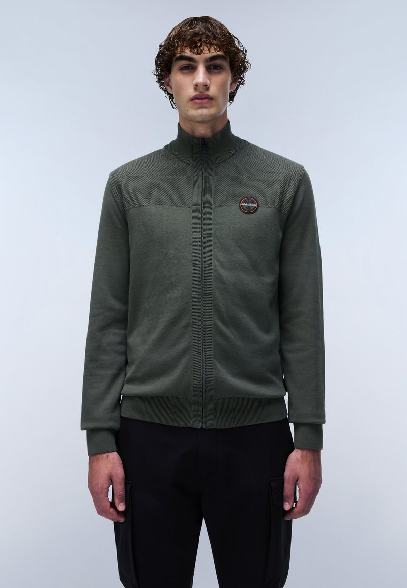 Un pull zippé vert avec un col montant, un panneau supérieur texturé et un patch de logo circulaire. Manches et ourlet côtelés, porté sur un pantalon sombre.