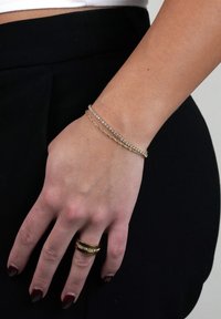 Guldtonad armband med flera strängar av klara rhinestones, bärs på ett handled, tillsammans med en guldring mot mörka kläder.