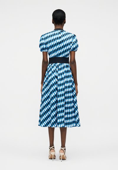 Γυναίκα με μαλλιά πιασμένα πίσω, που φορεί μεσομήκι φόρεμα με μπλε, μαύρο και λευκό μοτίβο, με φουσκωτά μανίκια και λευκά σανδάλια με τακούνι, με το πρόσωπο στραμμένο προς τα πίσω.